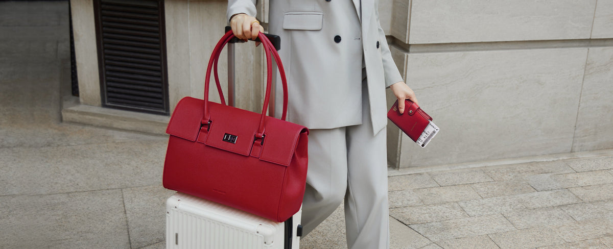 Professional woman in a grey suit carrying a structured red work bag and rolling luggage — Victoria Hyde London Jolene in red, built for business travel