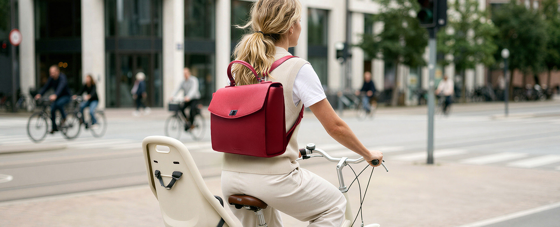 A business-dressed woman commuting by bicycle, equipped with an elegant red Potter backpack.