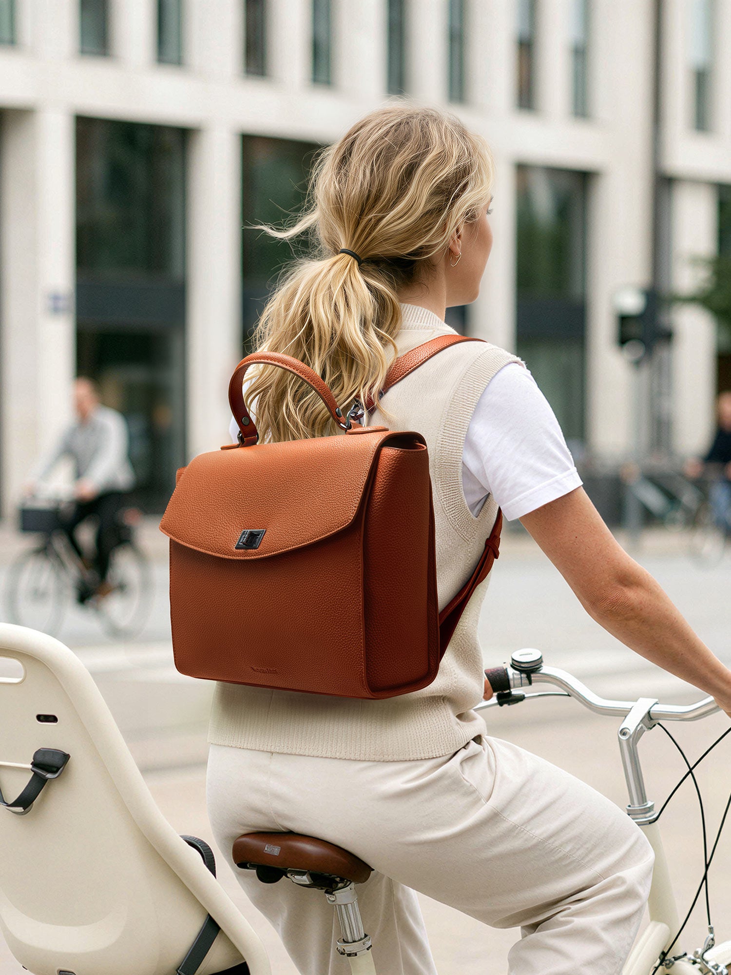 Woman cycling through the city wearing the Victoria Hyde Potter brown 2-in-1 convertible laptop backpack, showcasing its stylish vegan leather design for active professional commuters.